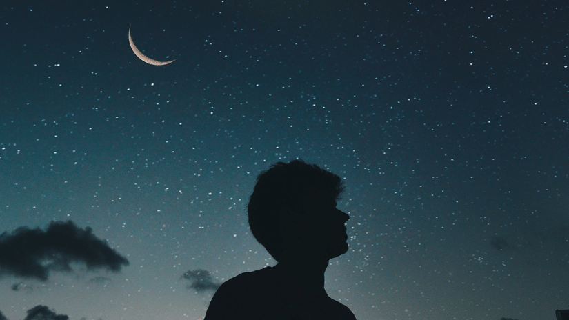 A silhouetted figure stands looking out at a dark night sky with a crescent moon and distant lights, possibly stars or city lights