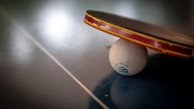 Table tennis paddle poised above a white ball on a dark playing surface, suggesting an impending serve or shot in a game