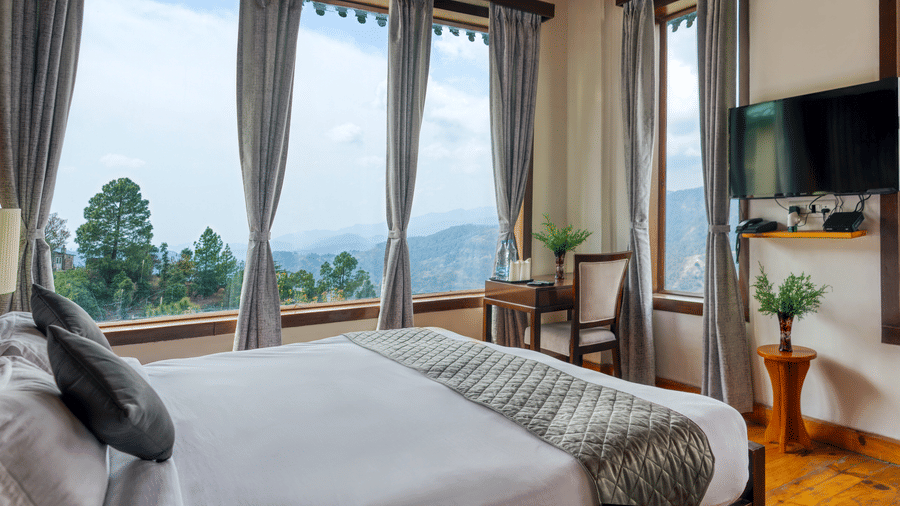 A comfortable hotel bedroom at Digantaa Resort in Mukteshwar featuring a large bed, a view of mountains through the window, and a television